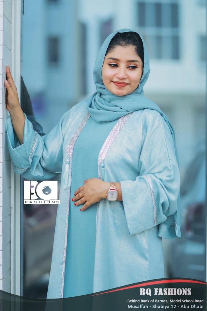 Young woman modeling light blue overcoat abaya with white lace trim detailing and matching blue hijab, standing indoors by glass entrance